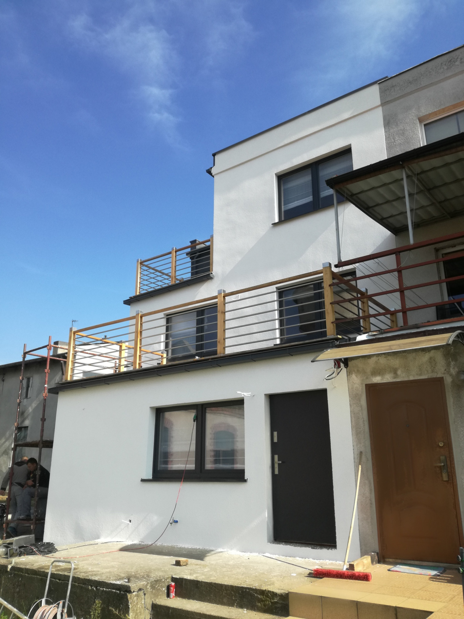 Dwupiętrowy dom z białymi ścianami i drewnianymi balustradami balkonów, widoczni pracownicy na rusztowaniu po lewej stronie, sugerujący trwające prace wykończeniowe.