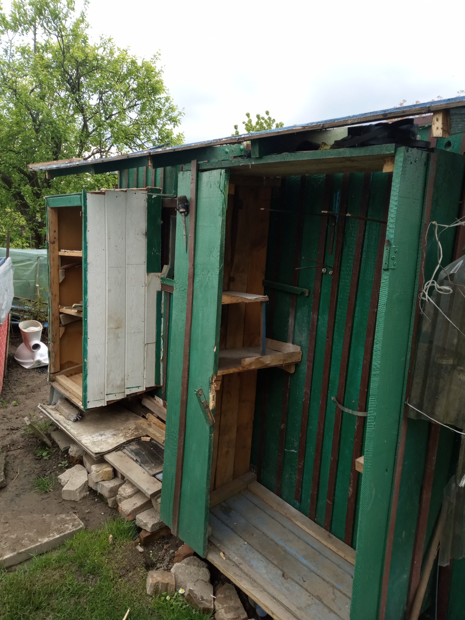 Niedokończona, prowizoryczna konstrukcja z drewna pomalowanego na zielono, z widocznymi półkami i fragmentami mebli, otoczona gruzem i elementami instalacji sanitarnych.