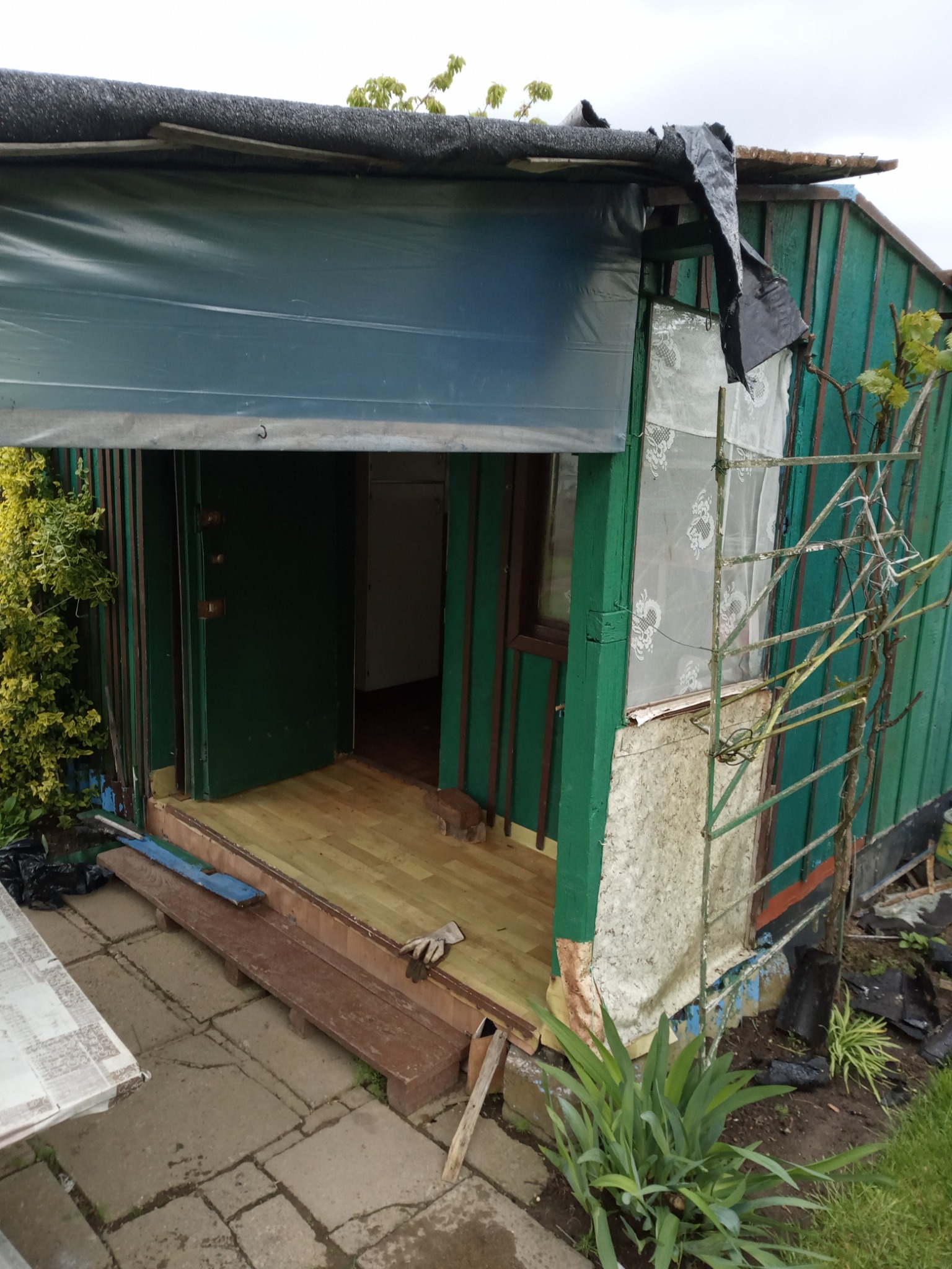 Niedokończona altana ogrodowa z zielonymi ścianami, prowizorycznym dachem z plandeki i drewnianą podłogą, widoczne narzędzia i fragmenty materiałów budowlanych w otoczeniu.