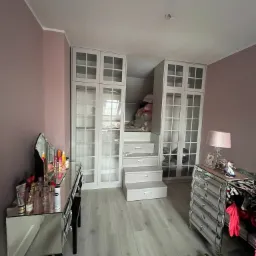 Pokój z antresolą i szafami w zabudowie, widoczne schody z szufladami prowadzące na antresolę, lustrzana toaletka, komoda i narzędzia na podłodze.