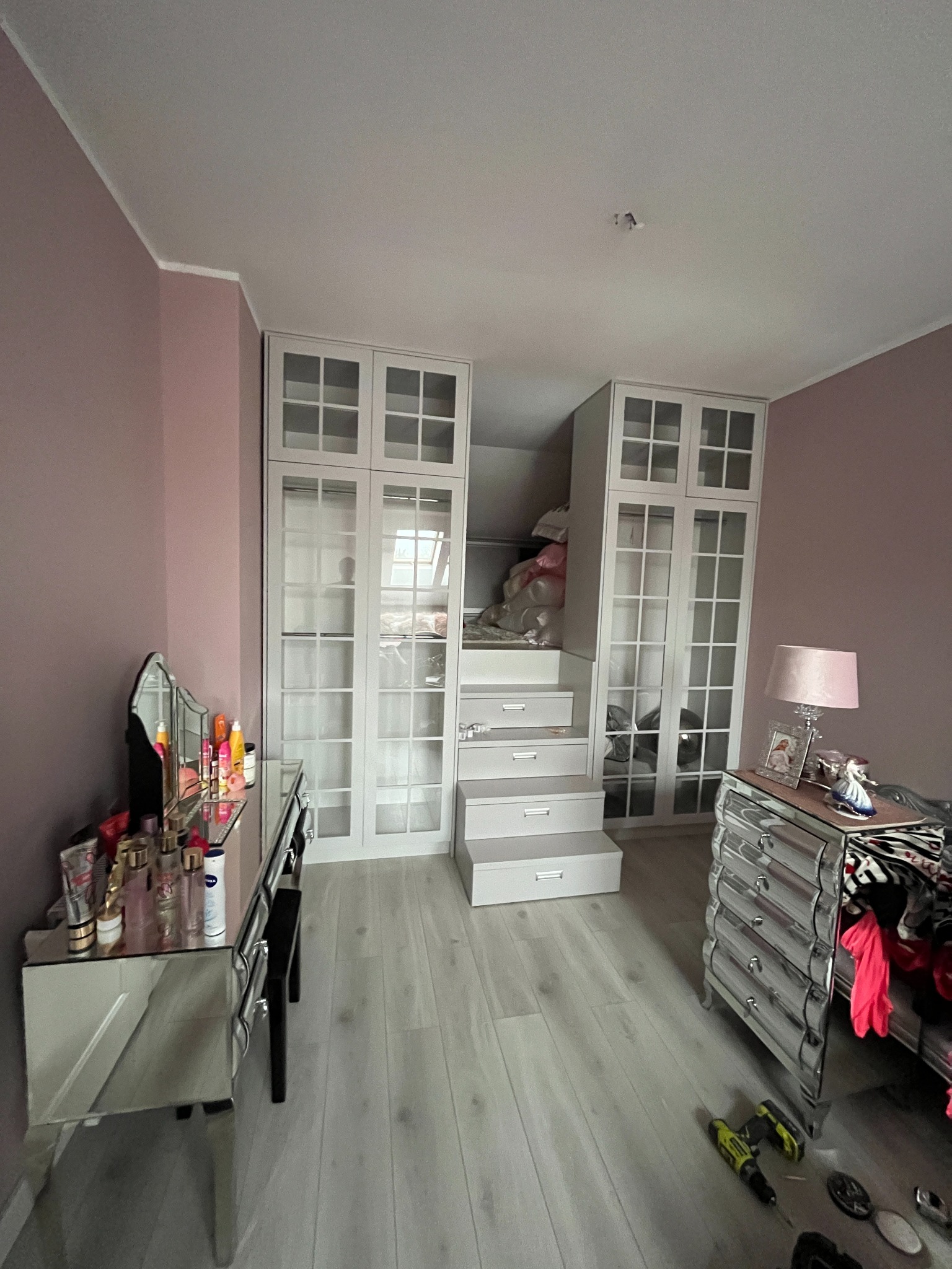 Pokój z antresolą i szafami w zabudowie, widoczne schody z szufladami prowadzące na antresolę, lustrzana toaletka, komoda i narzędzia na podłodze.