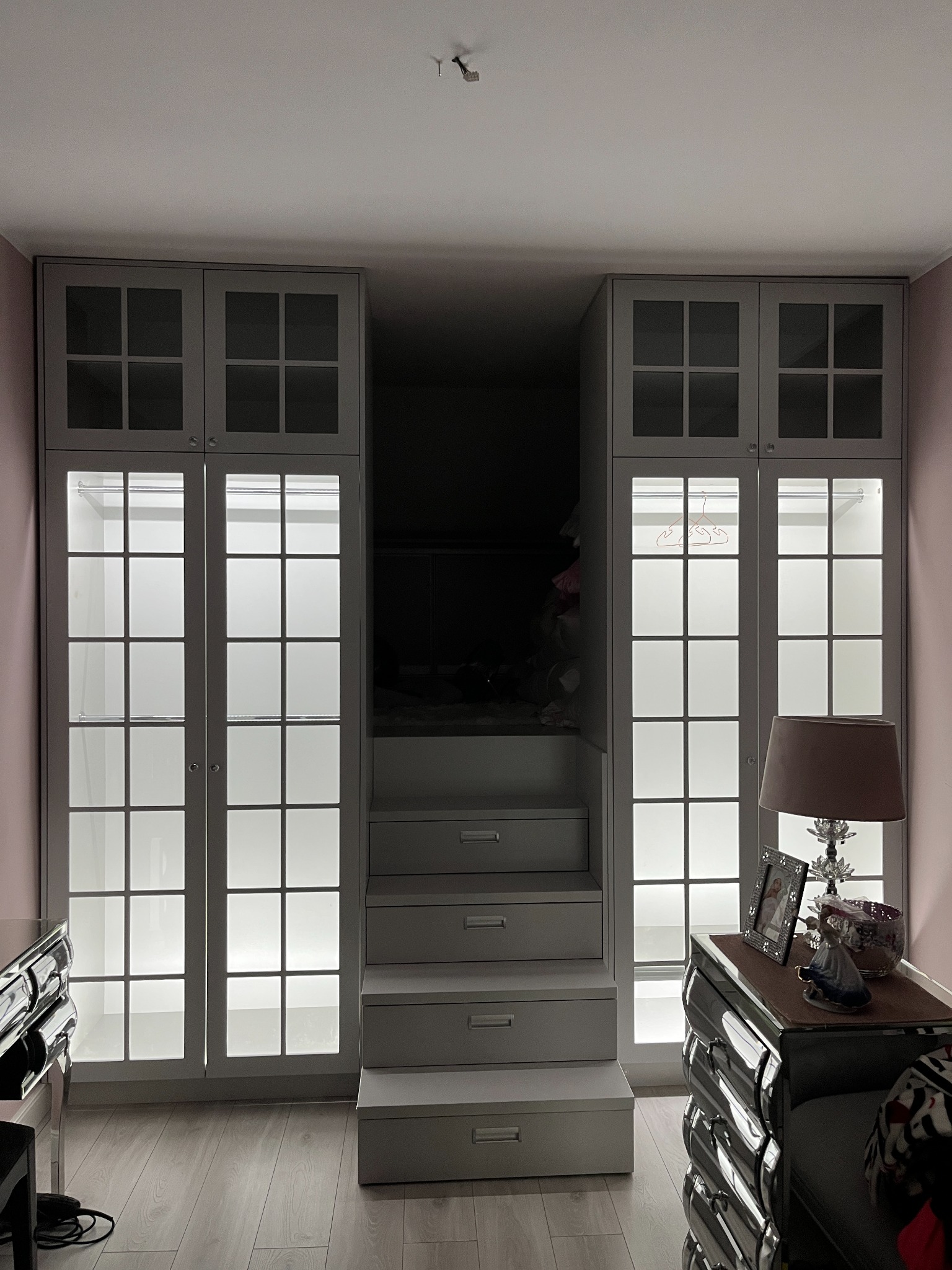 Szara zabudowa szafy z przeszklonymi drzwiami i wewnętrznym oświetleniem, zintegrowana ze schodami prowadzącymi do antresoli, widok na fragment szafki nocnej z lampką.