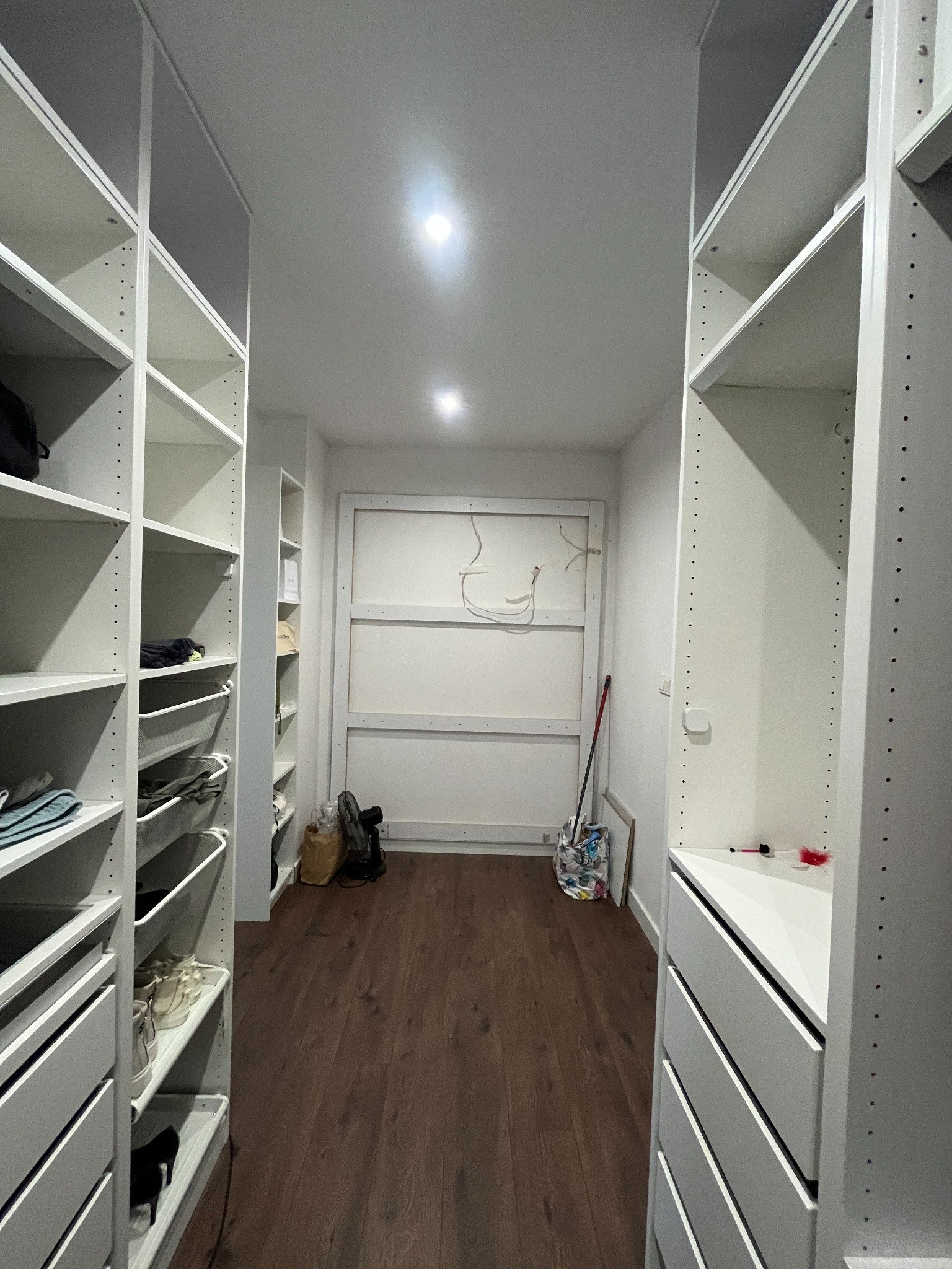 Wnętrze garderoby w trakcie montażu: białe regały z półkami i szufladami po bokach, widoczne ubrania i akcesoria, na podłodze narzędzia i akcesoria montażowe.