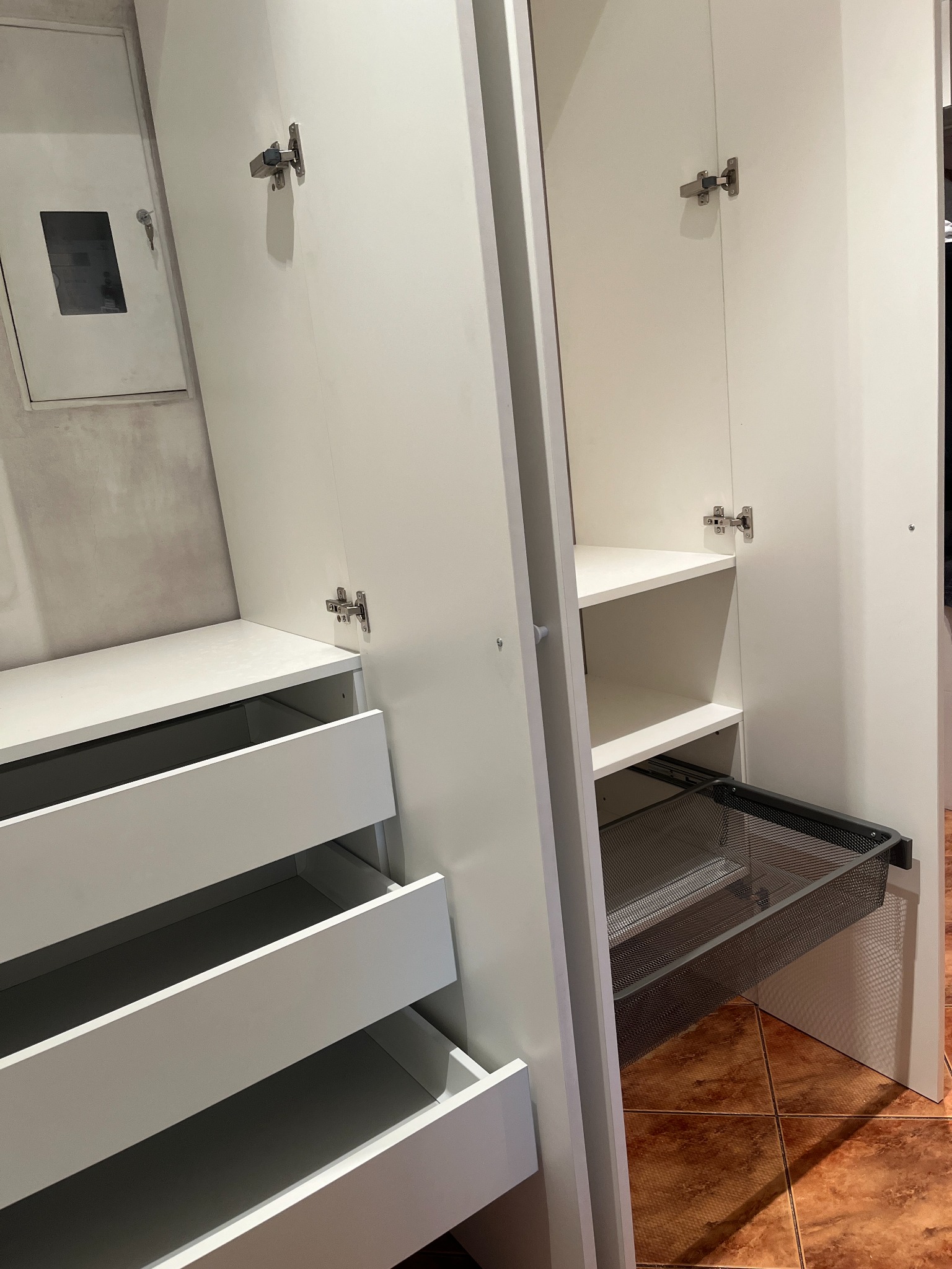 Wnętrze białej szafy z otwartymi drzwiami i wysuwanymi szufladami, widoczna metalowa siatkowa półka na prowadnicach, na ścianie obok wisi obrazek w białej ramie.