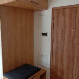 K & K MEBLE - Drewniana zabudowa przedpokoju z szafkami, siedziskiem i szufladami w jasnym odcieniu drewna, z czarnymi uchwytami, obok drzwi o podobnym wykończeniu.