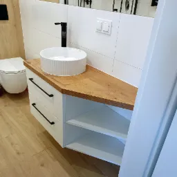 Nowoczesna łazienka: biała szafka z drewnianym blatem, okrągła umywalka i czarna bateria. Minimalistyczny design z płytkami i drewnianą podłogą.