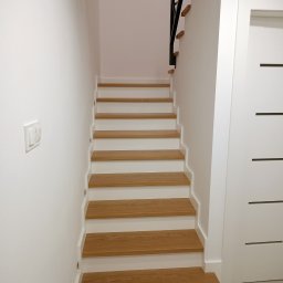 K & K MEBLE - Jasne drewniane schody z białymi podstopnicami i czarną balustradą, prowadzące w górę w minimalistycznym, białym wnętrzu. Obok widoczna biała ściana z włącznikiem i białe drzwi.