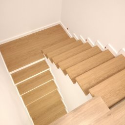 K & K MEBLE - Nowoczesne, drewniane schody z białymi podstopnicami, ujęcie z góry. Jasne drewno, minimalizm, proste linie. Widoczny fragment podłogi na dole schodów.