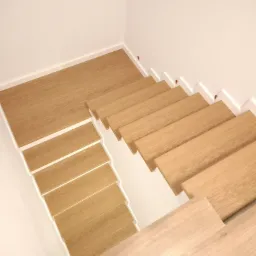Nowoczesne, drewniane schody z białymi podstopnicami, ujęcie z góry. Jasne drewno, minimalizm, proste linie. Widoczny fragment podłogi na dole schodów.