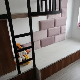 Wnętrze pokoju dziecięcego z antresolą: drewniane schody z czarną metalową ramą prowadzą na górę, obok znajduje się podest z szufladami i panelem tapicerowanym w kolorze pudrowego różu, na podeście...