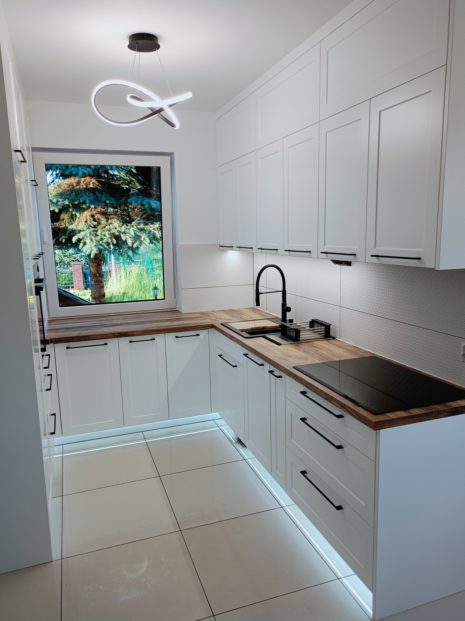 Biała kuchnia z drewnianym blatem, czarną armaturą i oświetleniem LED pod szafkami. Za oknem widok na zielony ogród. Nowoczesna lampa wisząca nad oknem.
