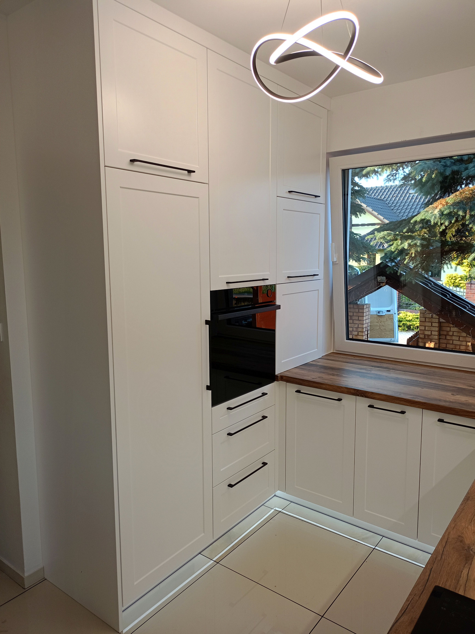 Nowoczesna, jasna kuchnia z białymi szafkami, czarnymi uchwytami i drewnianym blatem przy oknie. Widoczny piekarnik w zabudowie i designerska lampa.