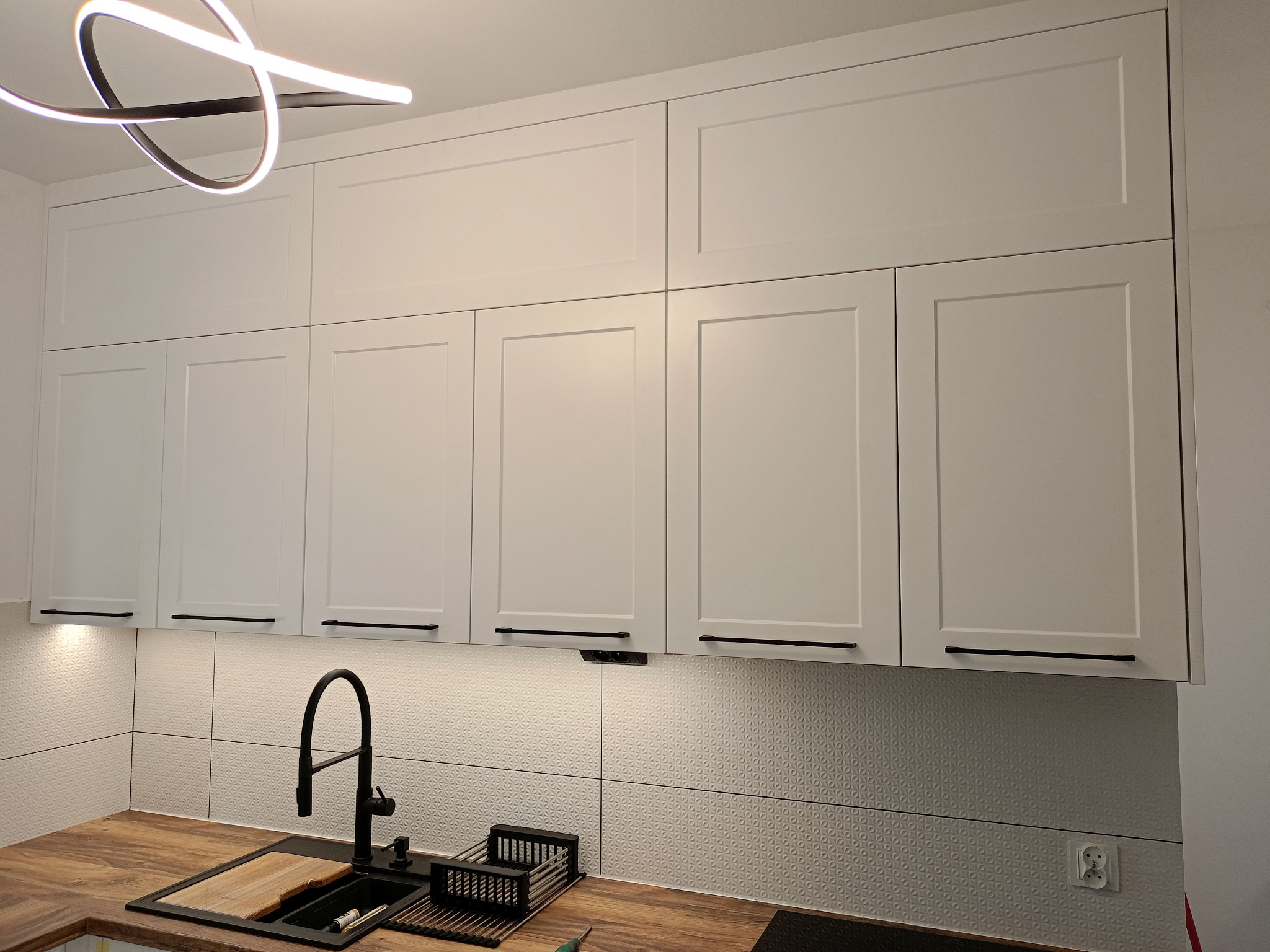 Nowoczesna kuchnia z białymi szafkami i drewnianym blatem. Czarna armatura i geometryczne płytki dodają elegancji. Oświetlenie LED podkreśla minimalistyczny design.