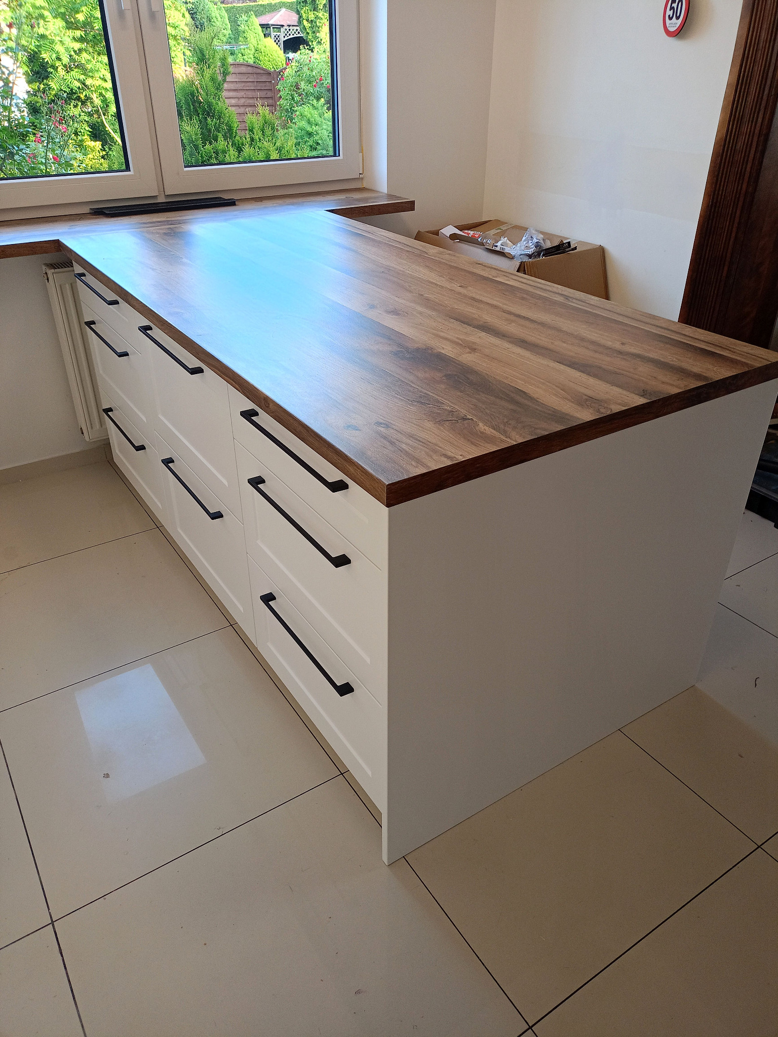 Wyspa kuchenna z białymi frontami i drewnianym blatem pod oknem. Długie, czarne uchwyty. Widoczne płytki podłogowe i fragment ściany z elementem dekoracyjnym.