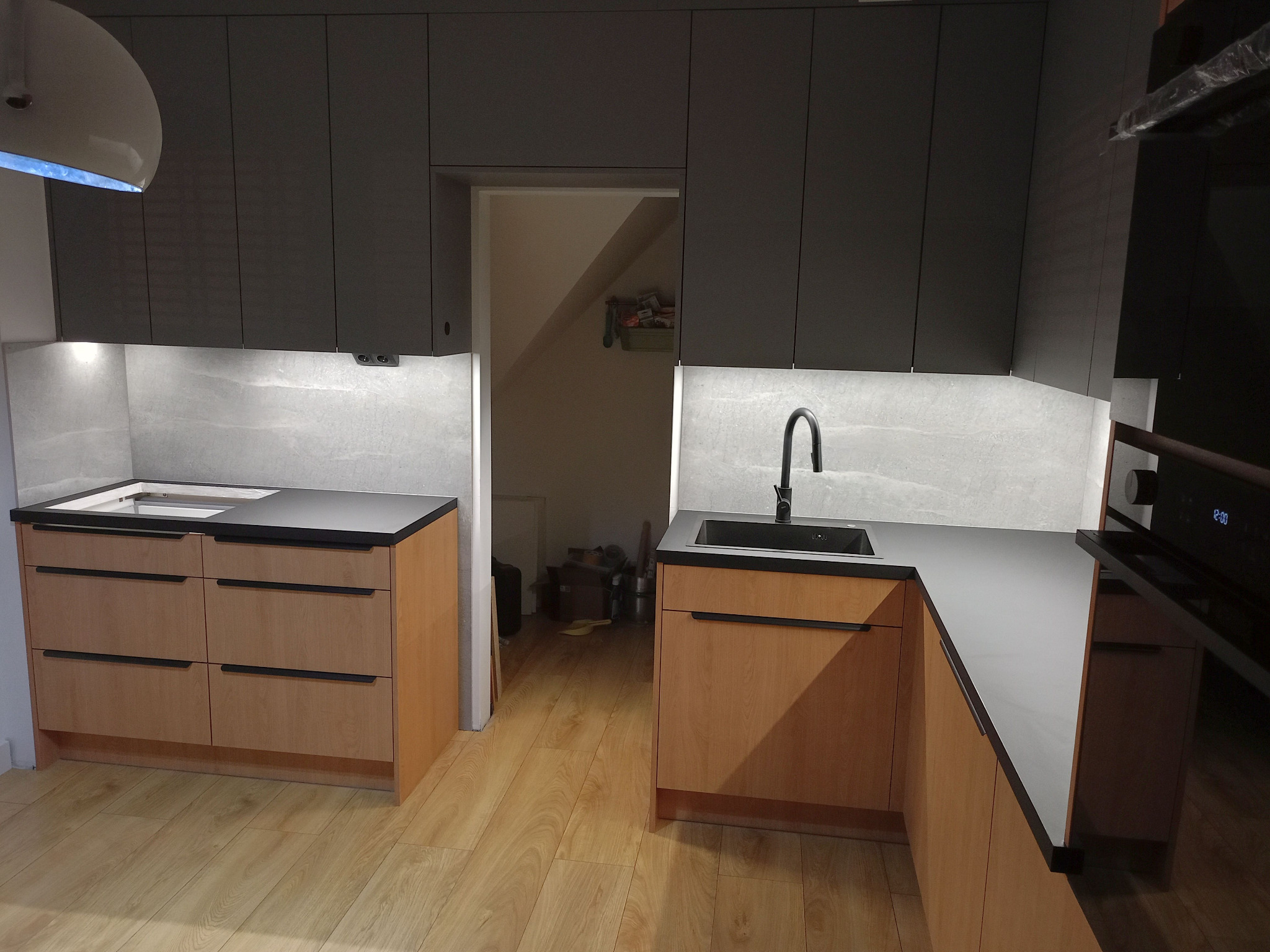 Nowoczesna kuchnia z drewnianymi frontami i szarymi szafkami wiszącymi. Blat w kolorze antracytowym, oświetlenie LED podszafkowe, drewniana podłoga, widok na pomieszczenie obok.