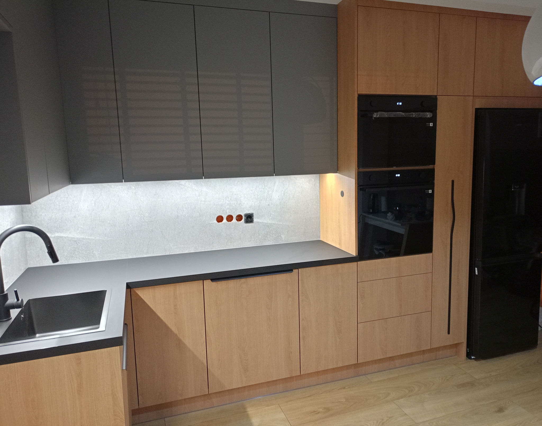 Nowoczesna kuchnia z szarymi szafkami górnymi i drewnopodobnymi dolnymi, czarnym blatem i sprzętem AGD, oświetlona listwą LED pod szafkami.