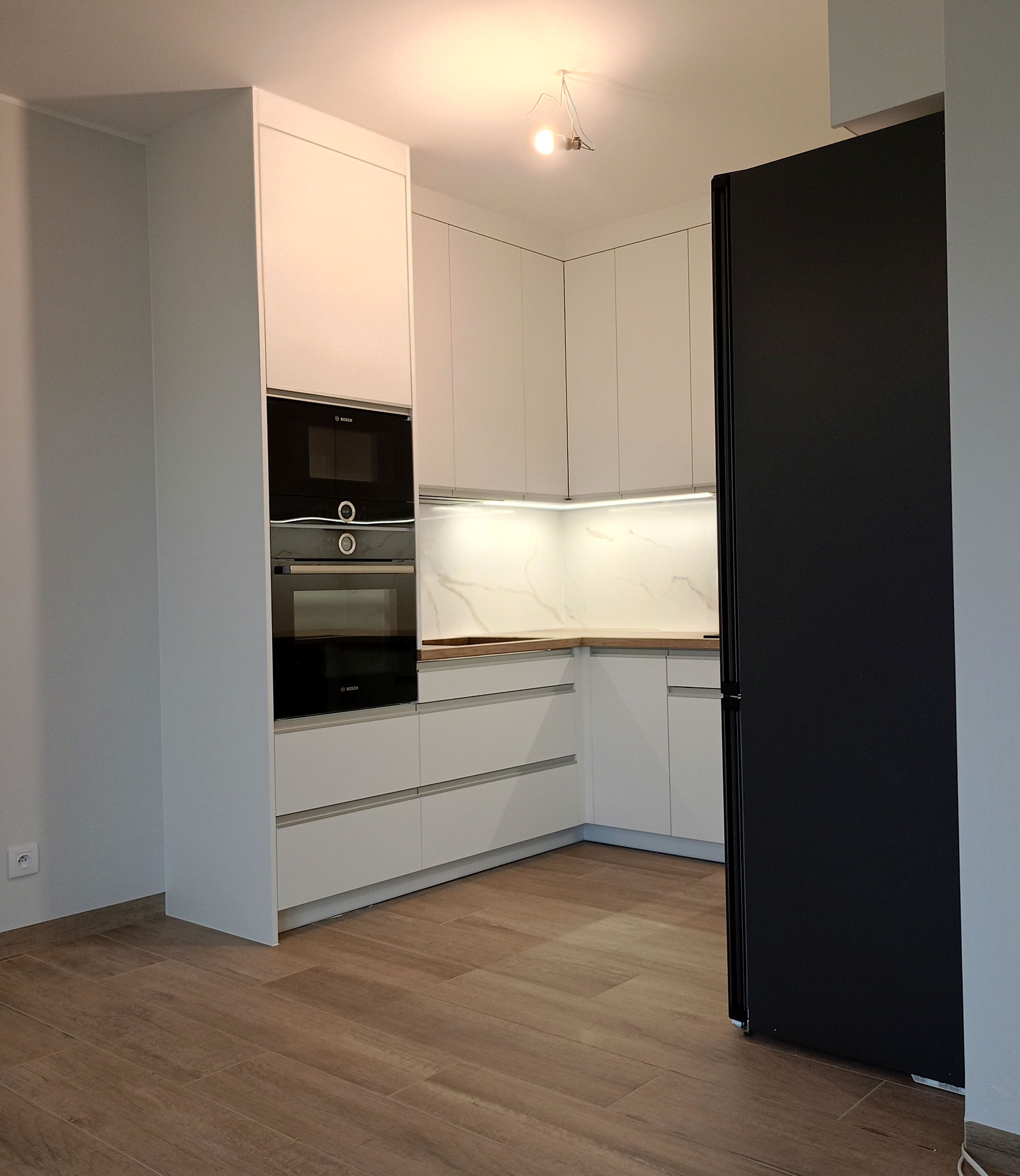 Nowoczesna, biała kuchnia z drewnianym blatem i czarnym piekarnikiem marki Bosch. Minimalistyczny design, oświetlenie LED pod szafkami, drewnopodobne płytki na podłodze.