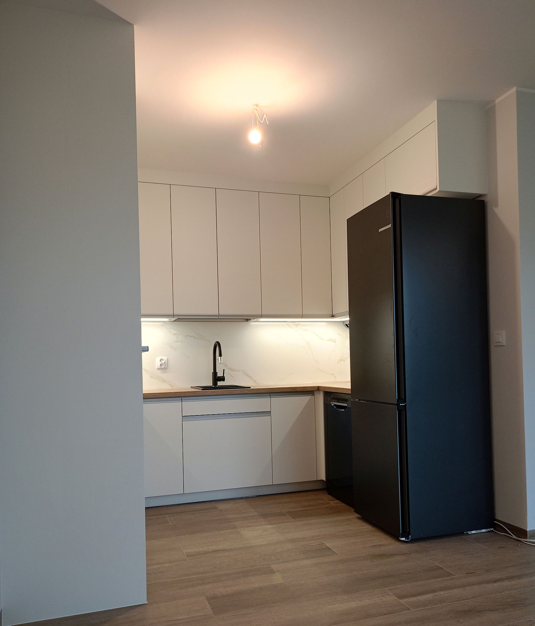 Nowoczesna kuchnia: białe szafki z drewnianym blatem, czarna armatura i lodówka. Minimalistyczny design, podświetlenie LED, płytki imitujące drewno na podłodze.