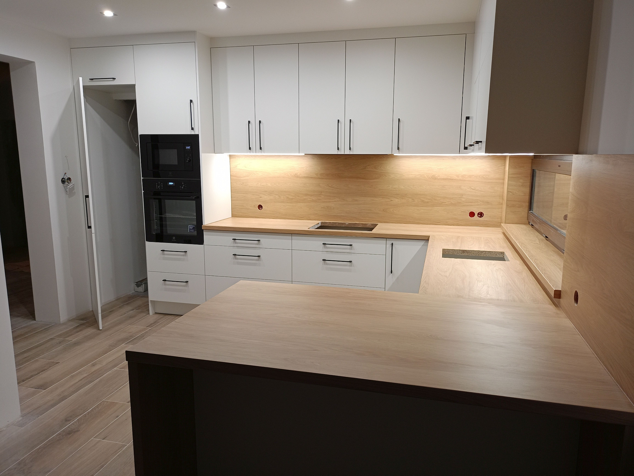 Nowoczesna kuchnia na wymiar w Chojnicach: białe szafki z czarnymi uchwytami, drewniany blat i oświetlenie podszafkowe. Minimalistyczny design.