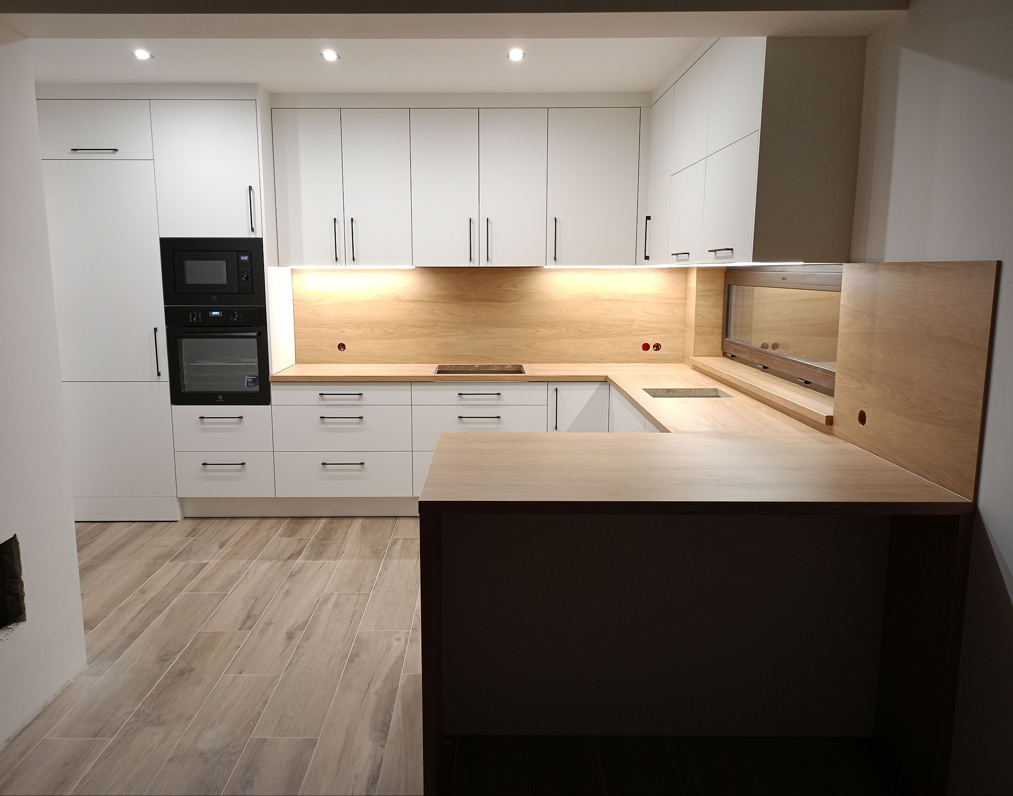 Nowoczesna, jasna kuchnia z białymi frontami i drewnianym blatem. Widoczne zabudowane sprzęty i oświetlenie podszafkowe. Uwagę przykuwa drewniana zabudowa okna.