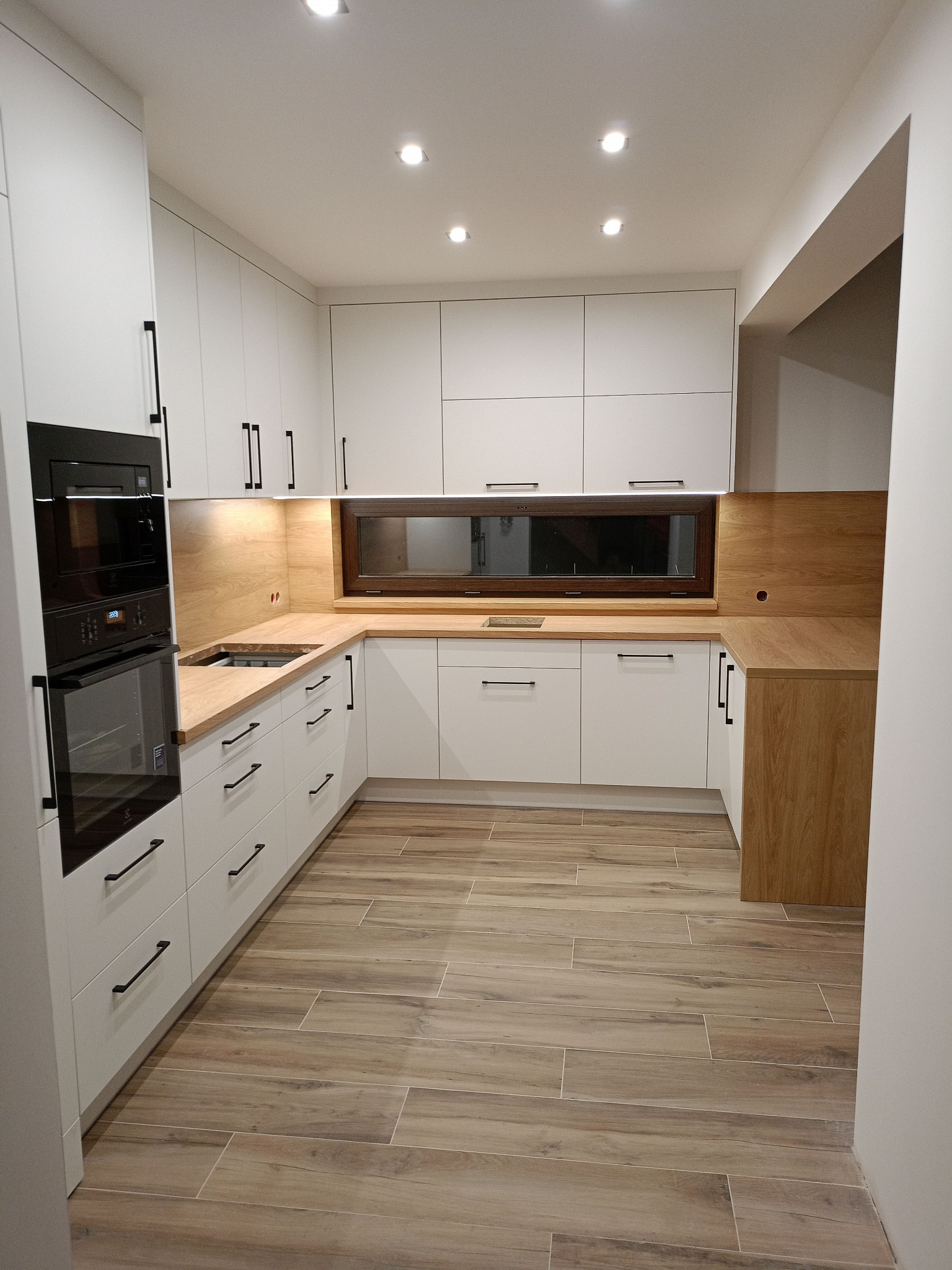 Jasna kuchnia na wymiar z drewnianym blatem, czarnymi uchwytami i oknem nad zlewem. Płytki podłogowe imitują drewno. Widoczny piekarnik w zabudowie.