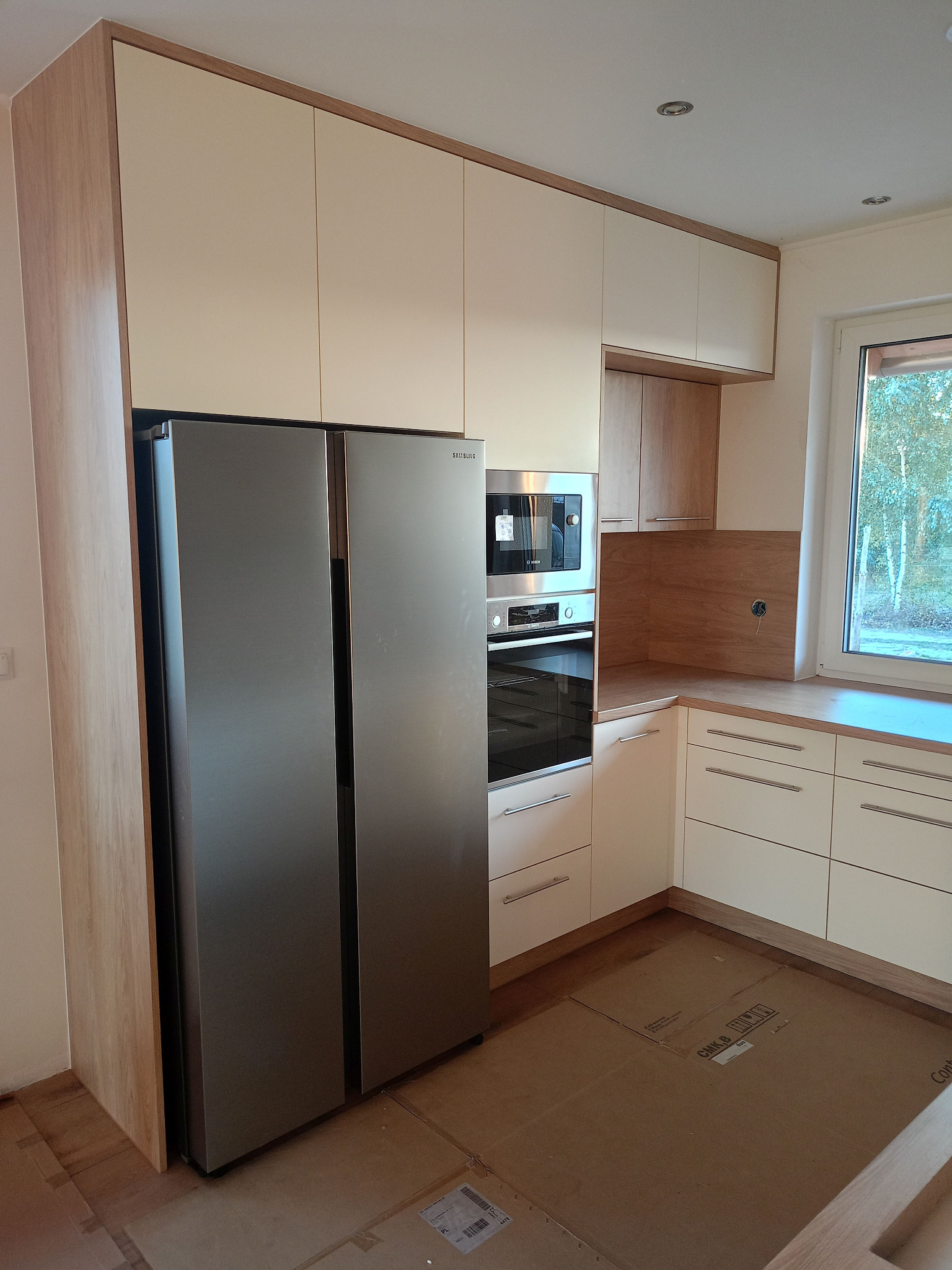 Jasna kuchnia z drewnianymi akcentami: zabudowa z szafkami górnymi i dolnymi, sprzęt AGD w zabudowie, blat z widocznym usłojeniem drewna, podłoga zabezpieczona kartonem.