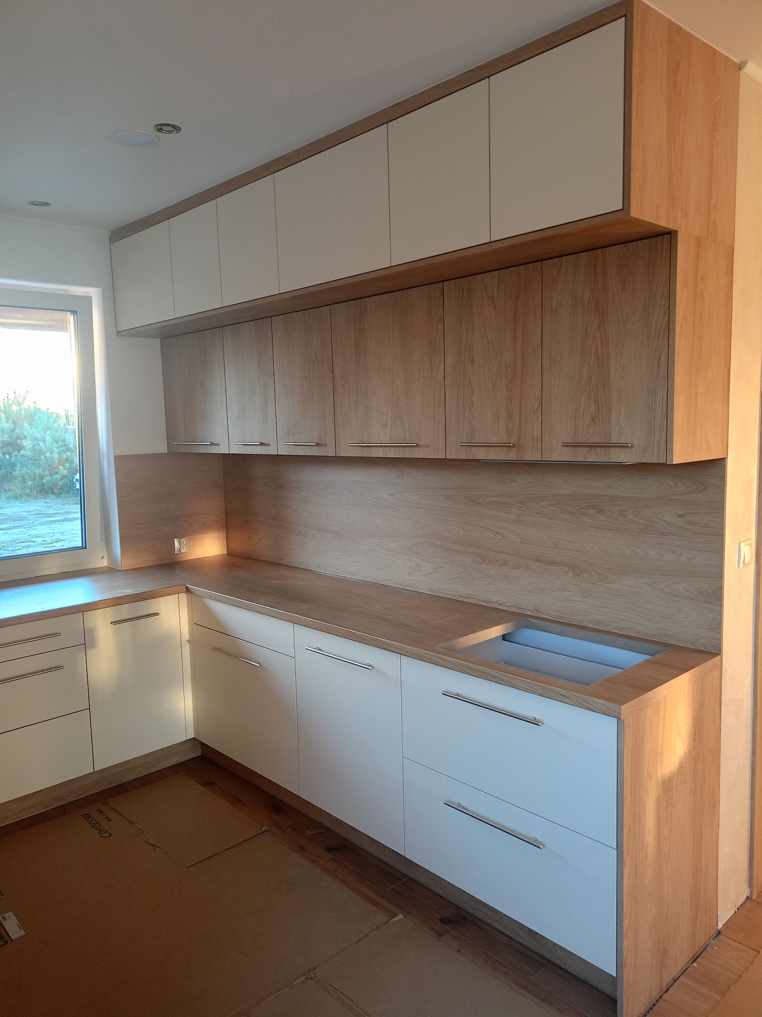 Jasna kuchnia z drewnianymi akcentami: białe fronty szafek dolnych, drewniane blaty i górne szafki, widok na okno. Oświetlenie podkreśla ciepło drewna.