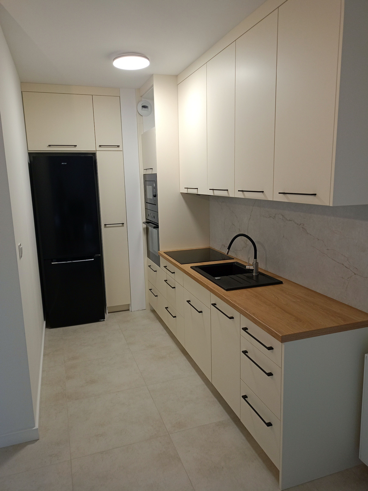 Beżowa kuchnia z drewnianym blatem i czarnymi uchwytami. W tle marmurowa ściana, czarny zlew i lodówka. Nowoczesne wykończenie, jasne płytki podłogowe.