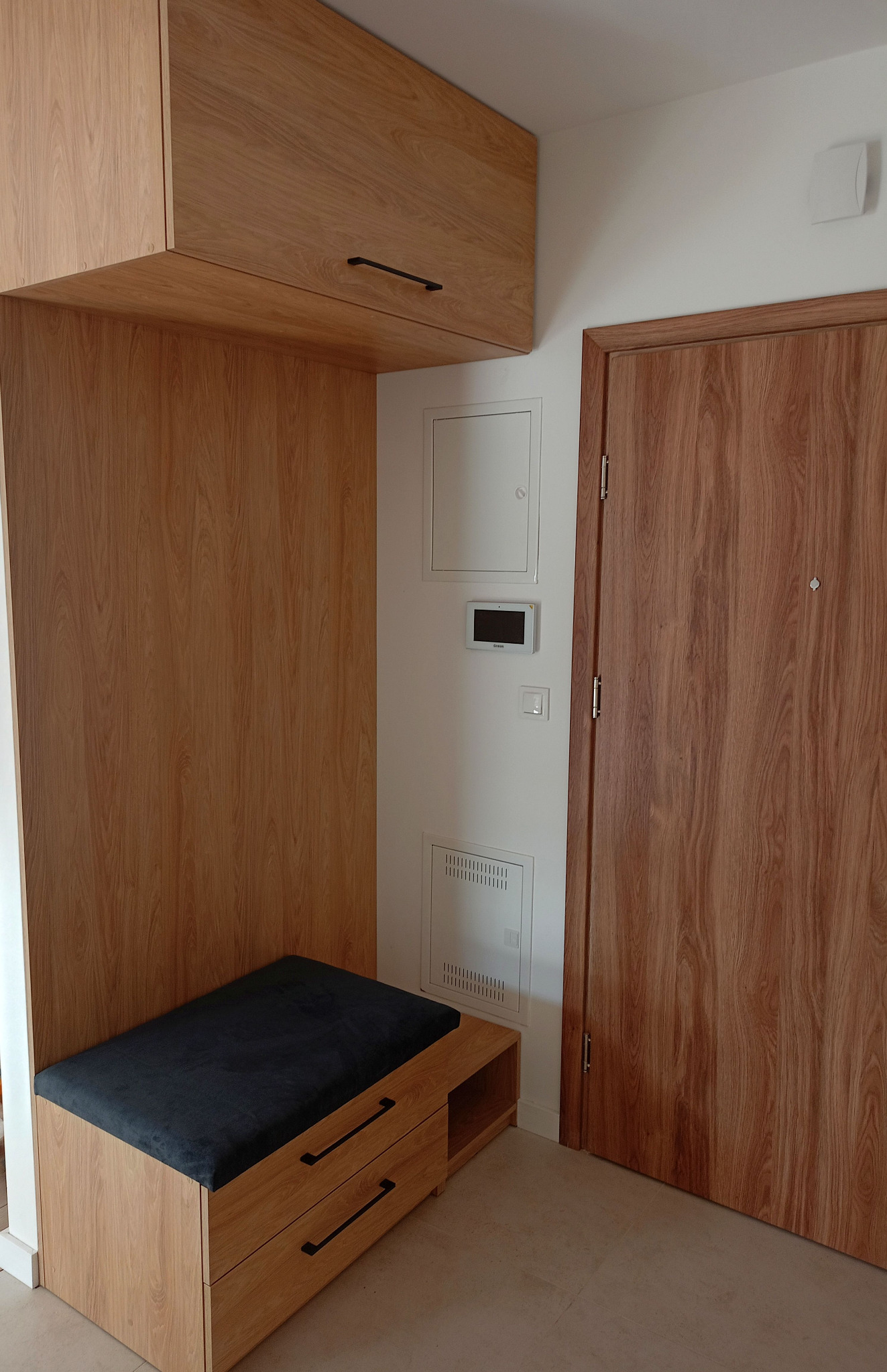 Drewniana zabudowa przedpokoju z szafkami, siedziskiem i szufladami w jasnym odcieniu drewna, z czarnymi uchwytami, obok drzwi o podobnym wykończeniu.