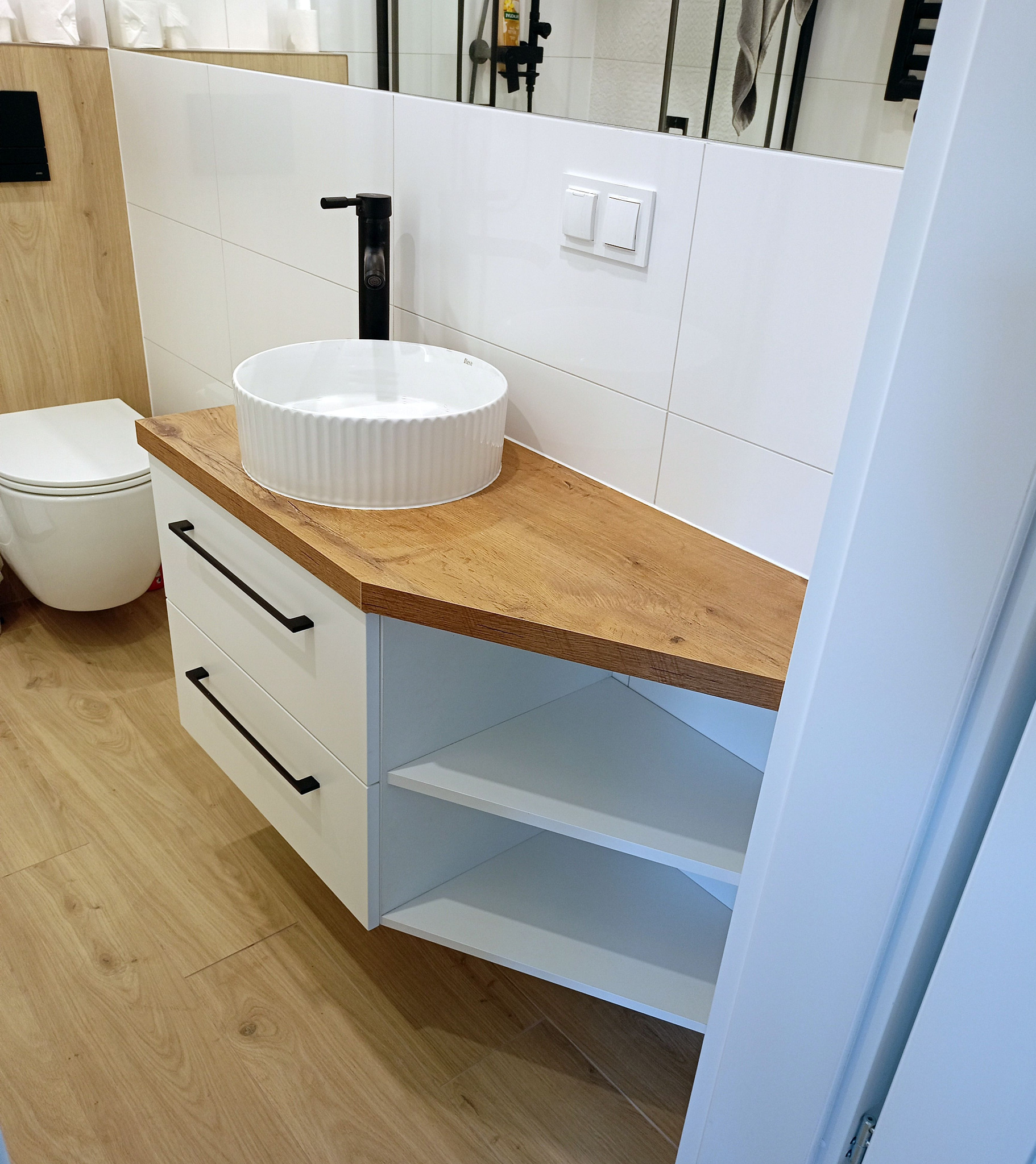 Nowoczesna łazienka: biała szafka z drewnianym blatem, okrągła umywalka i czarna bateria. Minimalistyczny design z płytkami i drewnianą podłogą.