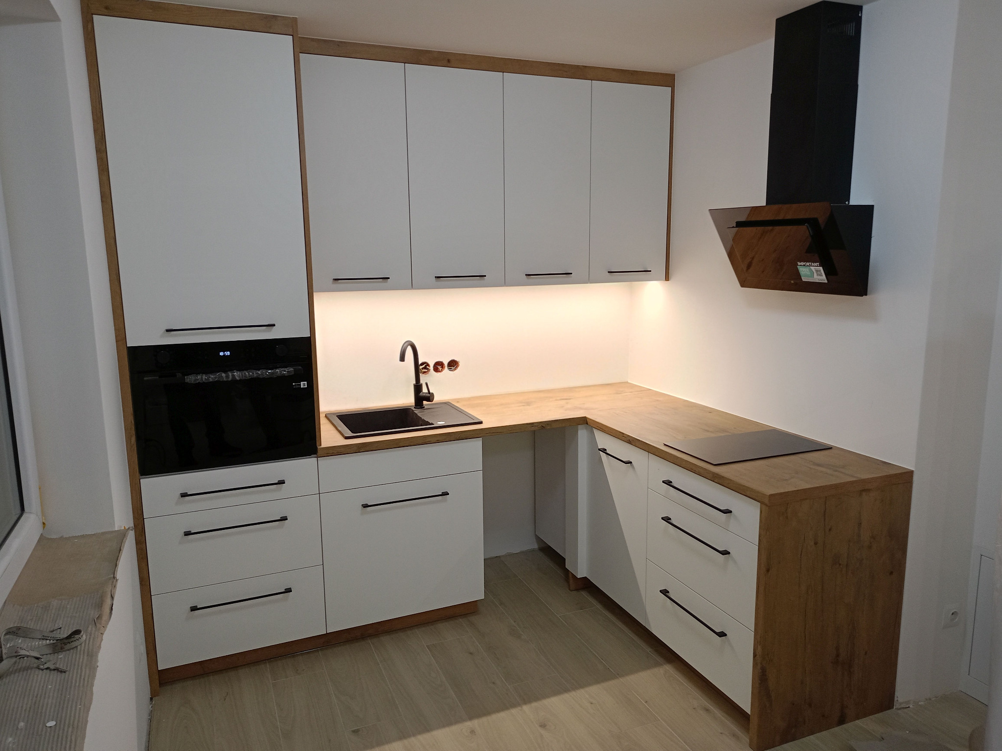 Nowoczesna kuchnia na wymiar z białymi frontami i drewnianym blatem. Czarny zlew i płyta indukcyjna. Ujęcie z perspektywy narożnika pomieszczenia.