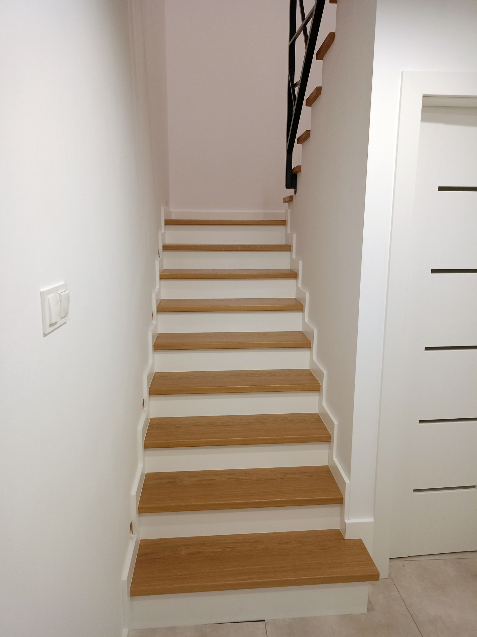 Jasne drewniane schody z białymi podstopnicami i czarną balustradą, prowadzące w górę w minimalistycznym, białym wnętrzu. Obok widoczna biała ściana z włącznikiem i białe drzwi.