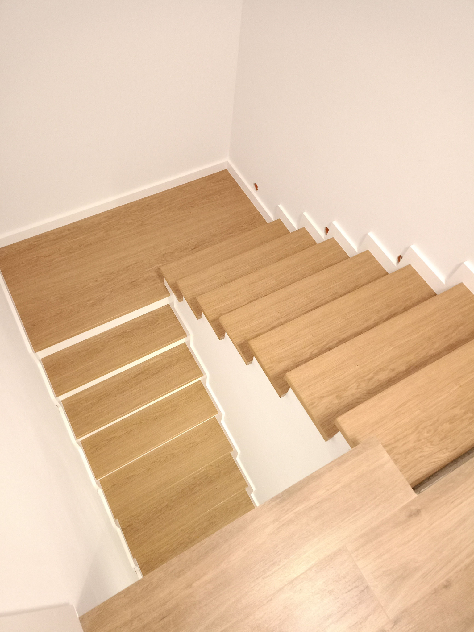 Nowoczesne, drewniane schody z białymi podstopnicami, ujęcie z góry. Jasne drewno, minimalizm, proste linie. Widoczny fragment podłogi na dole schodów.