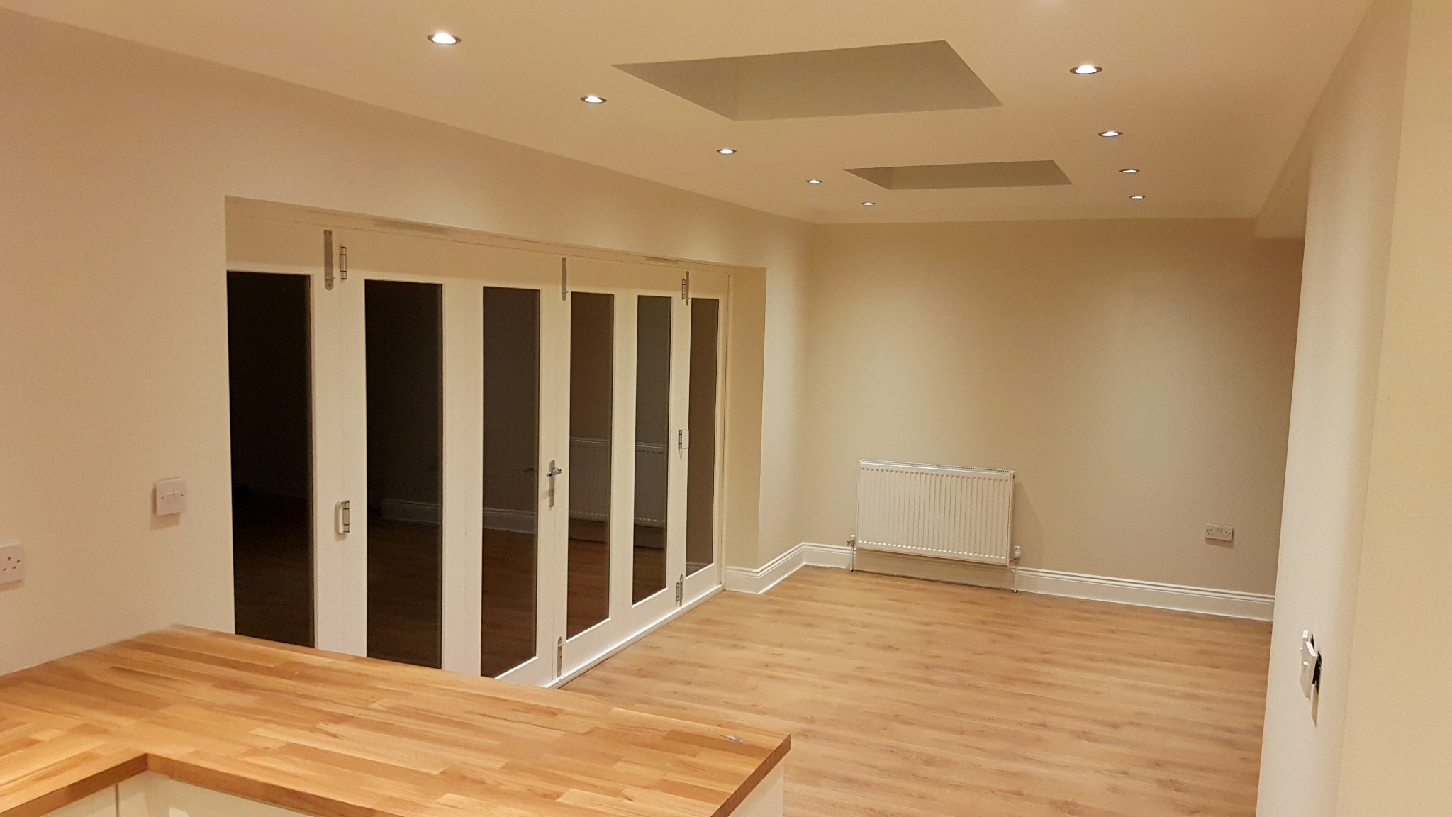 Jasne wnętrze domu szkieletowego z drewnianą podłogą, białymi ścianami i składanymi szklanymi drzwiami w stylu skandynawskim, widoczne grzejniki i oświetlenie sufitowe.