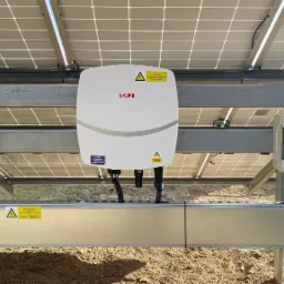 Instalacja fotowoltaiczna: widok na inwerter SAJ i dwie rozdzielnice PV-DC oraz PV-AC, zamocowane na metalowej konstrukcji pod panelami słonecznymi, z widocznymi przewodami i naklejkami...