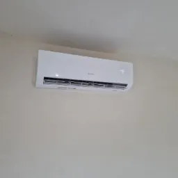 Biały klimatyzator marki Gree zamontowany na beżowej ścianie w pomieszczeniu, z widocznym wyświetlaczem temperatury i fragmentem okna w prawym dolnym rogu.