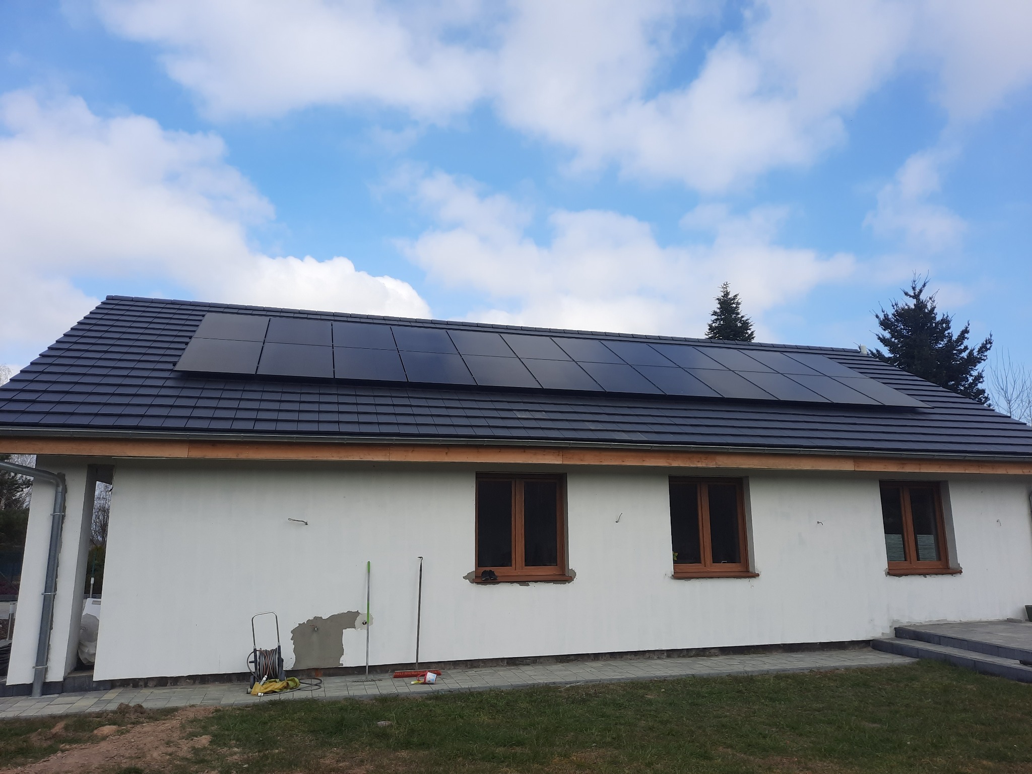 Panele słoneczne na dachu domu jednorodzinnego z białą elewacją, widoczne ślady prac wykończeniowych na ścianie i narzędzia ogrodowe obok budynku, błękitne niebo z chmurami w tle.