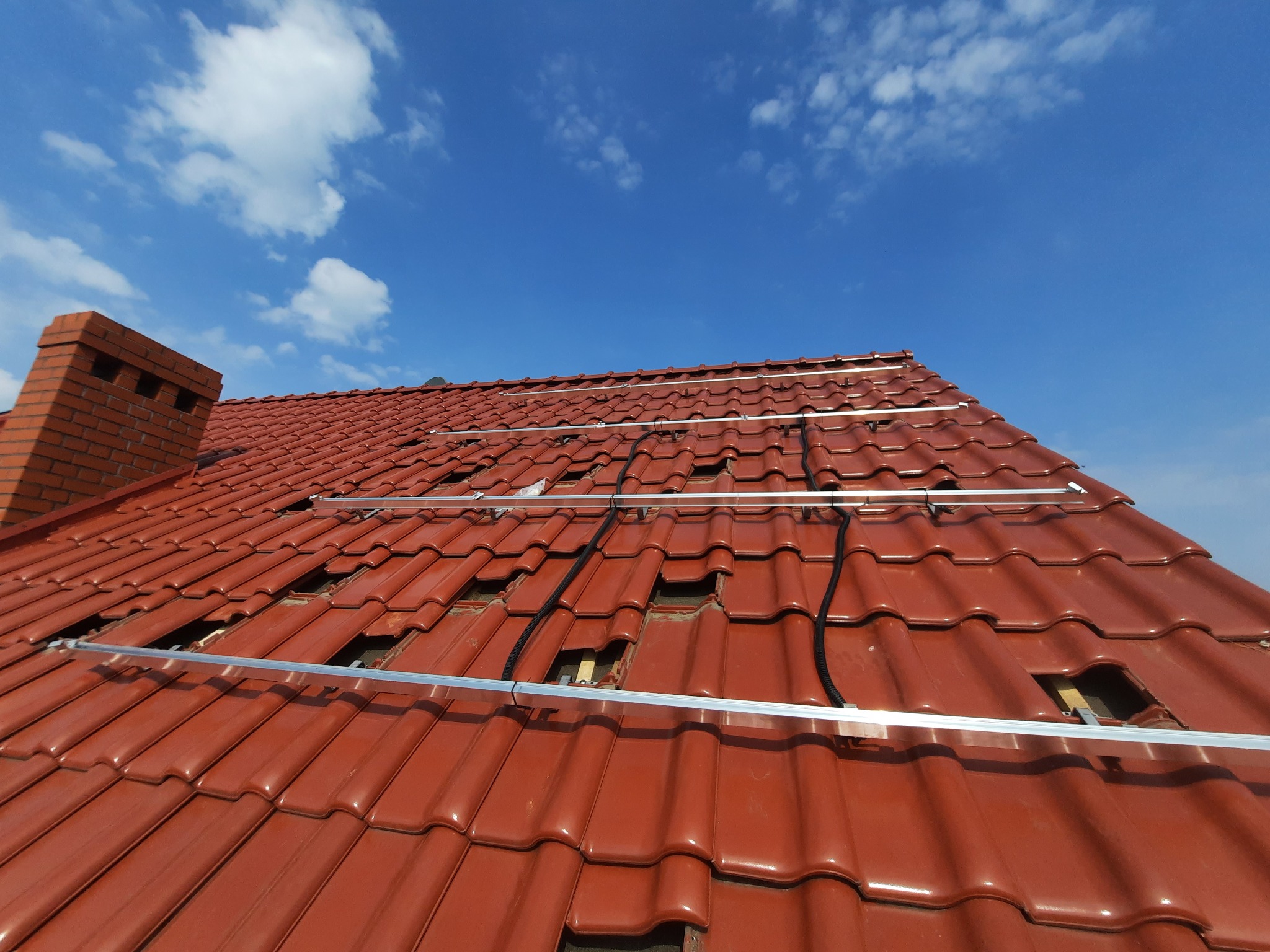 Montaż szyn pod panele fotowoltaiczne na dachu z czerwonej dachówki ceramicznej, widoczne kable, komin z cegły i błękitne niebo z chmurami w tle.