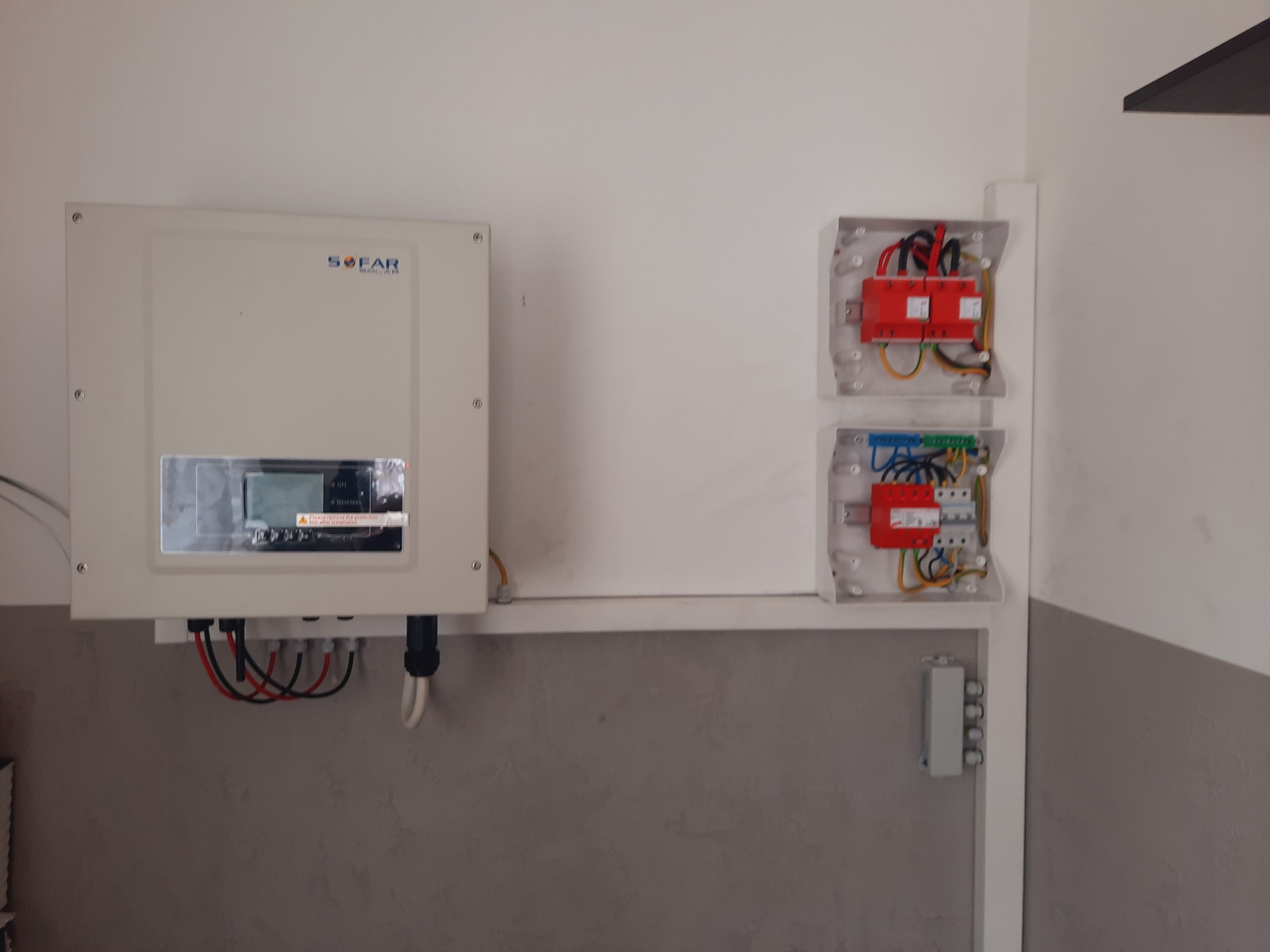 Biały falownik SOFAR zamontowany na ścianie nad szarą lamperią, obok dwie otwarte skrzynki z zabezpieczeniami i okablowaniem.
