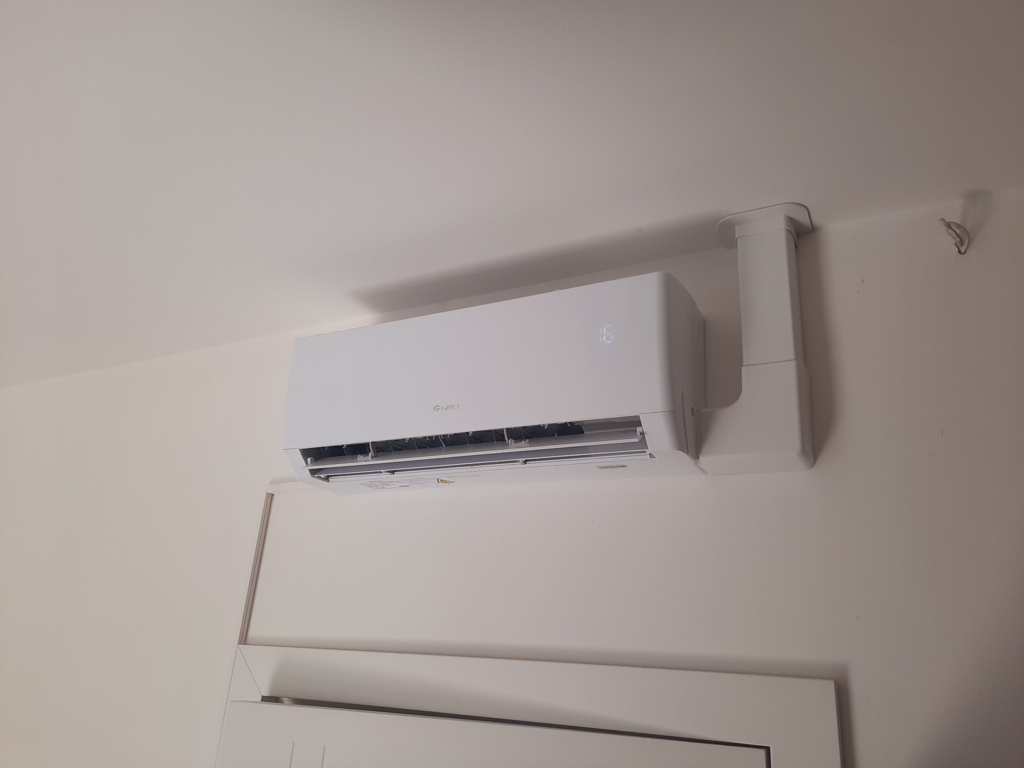 Biały klimatyzator marki Gree zamontowany na ścianie, widoczny fragment drzwi poniżej. Na wyświetlaczu jednostki wewnętrznej widoczna temperatura 16 stopni.