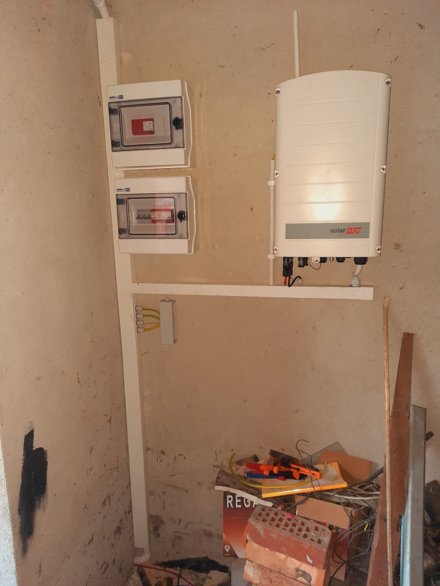 Zamontowany inwerter SolarEdge na ścianie w pomieszczeniu z dwoma skrzynkami bezpiecznikowymi, widoczne przewody i fragment nieporządku na podłodze.