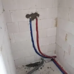 Wewnętrzny narożnik pomieszczenia w trakcie prac instalacyjnych: widoczne nieotynkowane ściany z białego bloczka, wystające z otworu w ścianie rury w izolacji czerwonej i niebieskiej, oraz szara...