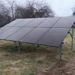 Czarny panel fotowoltaiczny zamontowany na metalowej konstrukcji na trawniku, widoczne drzewa bez liści w tle i trampolina.