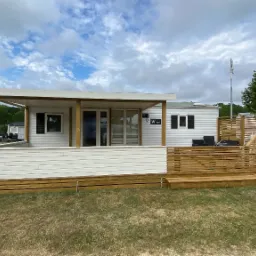 Nowoczesny, biały dom modułowy z drewnianym tarasem i ażurową osłoną, widok z zewnątrz w słoneczny dzień.