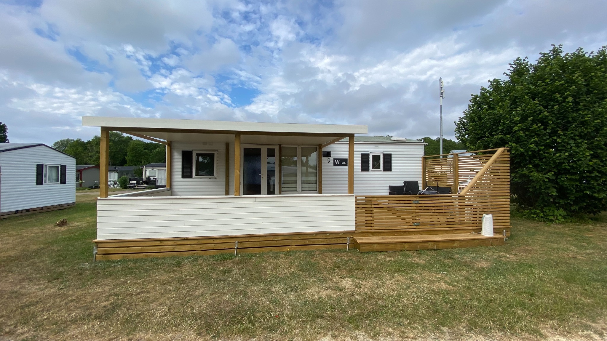 Nowoczesny, biały dom modułowy z drewnianym tarasem i ażurową osłoną, widok z zewnątrz w słoneczny dzień.