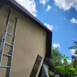 Ściana budynku z beżowym tynkiem, brązowe obramowanie dachu, metalowa drabina oparta o ścianę, błękitne niebo z białymi chmurami w tle, drzwi na ścianie.