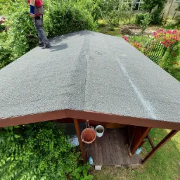 Widok z góry na pokryty papą dach altany ogrodowej, z fragmentem sylwetki dekarza w górnym lewym rogu, otoczonej zielenią.