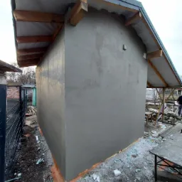 Świeżo otynkowana ściana budynku z widocznymi elementami drewnianej konstrukcji dachu, na ziemi pozostałości materiałów budowlanych.