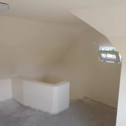 Pomalowane na kremowo poddasze z małym oknem, widoczne ślady po remoncie na podłodze.