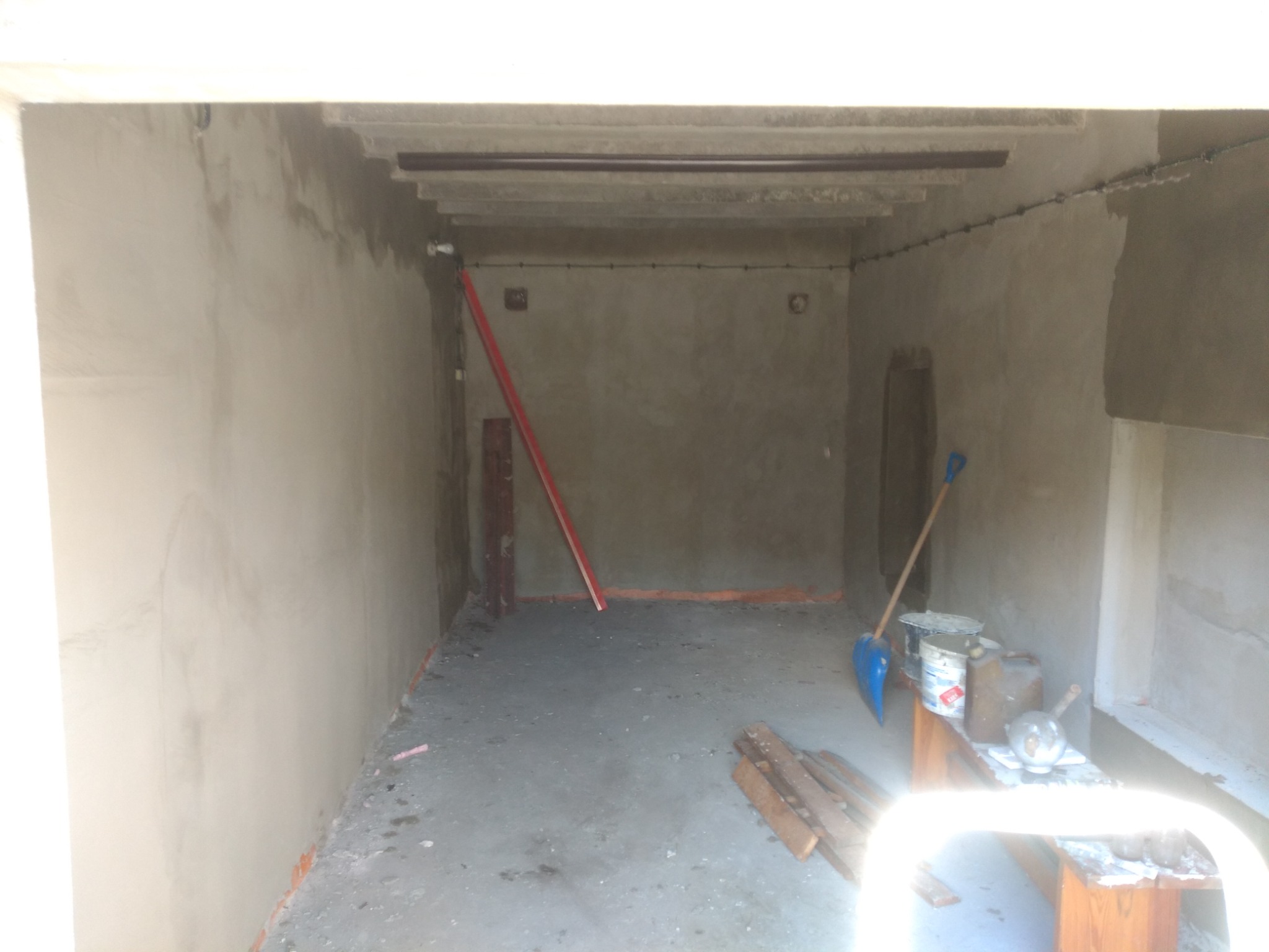 Surowe, betonowe wnętrze garażu w trakcie prac wykończeniowych; widoczne ślady świeżego tynku, narzędzia i materiały budowlane na drewnianym stole.